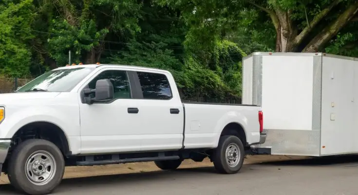 A white pickup truck pulls a white enclosed trailer. The truck and trailer are parked next to large trees.