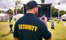 A man wearing a blue shirt that says "Security" standing in front of an outdoor stage while holding a two-way radio.