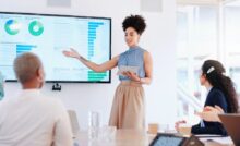 A woman is leading a seminar while holding a digital tablet. She's presenting stats on a large monitor.