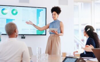 A woman is leading a seminar while holding a digital tablet. She's presenting stats on a large monitor.