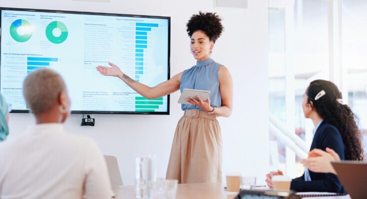 A woman is leading a seminar while holding a digital tablet. She's presenting stats on a large monitor.