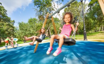 Two little girls are swinging on a swing with a black seat and chain links. The flooring is a blue rubber.