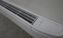 A close-up, top-down view of a white electric baseboard heater installed on a beige wall above gray flooring.
