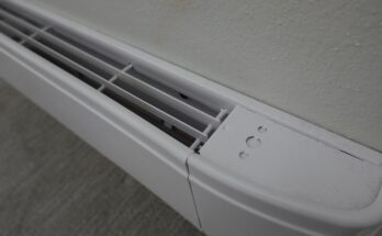 A close-up, top-down view of a white electric baseboard heater installed on a beige wall above gray flooring.