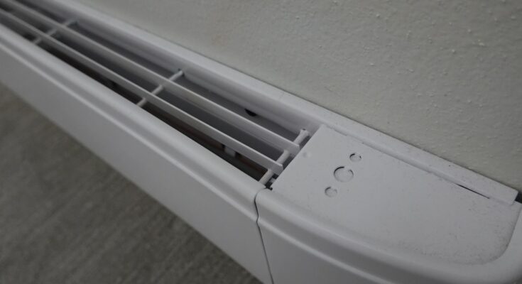 A close-up, top-down view of a white electric baseboard heater installed on a beige wall above gray flooring.