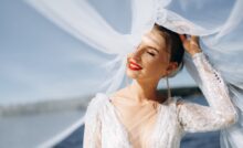 A joyful bride in soft sunlight, wearing a flowing veil and sparkling dress, stands near a body of water.