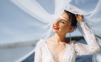 A joyful bride in soft sunlight, wearing a flowing veil and sparkling dress, stands near a body of water.