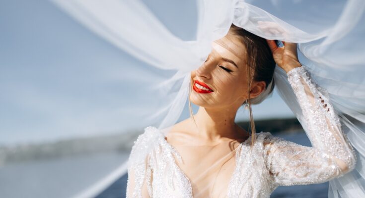 A joyful bride in soft sunlight, wearing a flowing veil and sparkling dress, stands near a body of water.