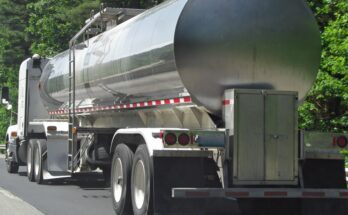 A silver tanker trailer is attached to a white truck. The truck and trailer sit on the side of the road.