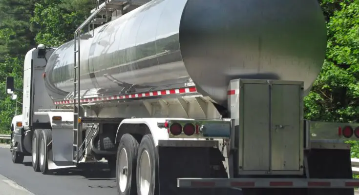 A silver tanker trailer is attached to a white truck. The truck and trailer sit on the side of the road.