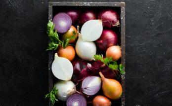 A selection of cut and whole onions sit in a black wooden box on a black countertop. Sprigs of parsley garnish the box.