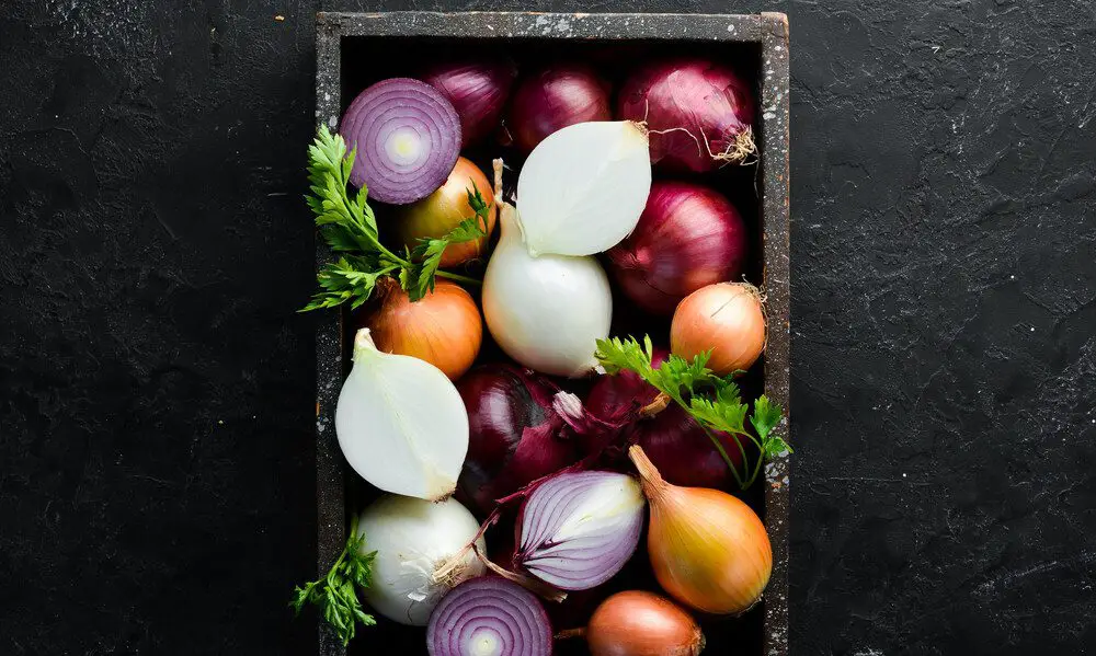 A selection of cut and whole onions sit in a black wooden box on a black countertop. Sprigs of parsley garnish the box.
