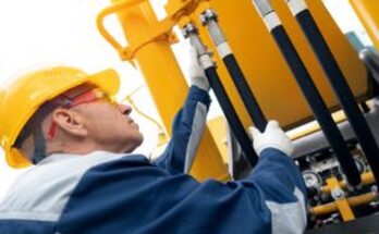 A man in a safety jacket, gloves, goggles, and hard hat inspects the hydraulic hose system on an excavator.