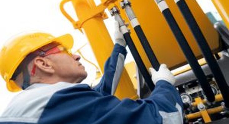 A man in a safety jacket, gloves, goggles, and hard hat inspects the hydraulic hose system on an excavator.