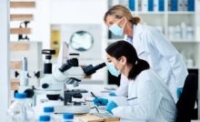 One female scientist sits in a lab looking through a black-and-white microscope. Another woman is looking at the table.