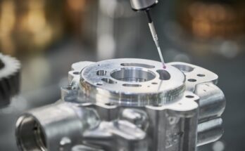 A close-up view shows a piece of silver machinery that is being measured by a stylus with a pink ball.