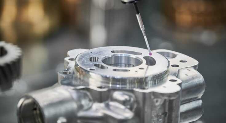 A close-up view shows a piece of silver machinery that is being measured by a stylus with a pink ball.
