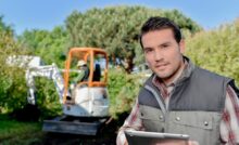 A man wearing a gray, puffy vest and a plaid shirt holds a tablet. A person operates a mini excavator behind him.