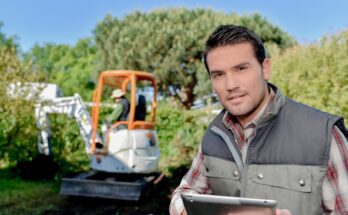 A man wearing a gray, puffy vest and a plaid shirt holds a tablet. A person operates a mini excavator behind him.