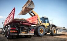A yellow excavator lifts up a pile of dirt to dump in the back of an orange-colored dump truck at a construction site.