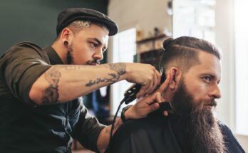 A man with a hat uses a black hair trimmer to trim the hair of a man with a long beard sitting in a chair.