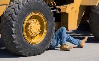 A person wearing jeans and brown work boots lies on the ground under a piece of large, yellow heavy machinery.