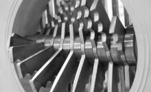 A close-up view shows a silver, metal industrial crusher. The rotating teeth have three grooves.