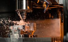 A close-up of a CNC machine drilling into a metal part as cutting fluid splashes onto and off of the part.