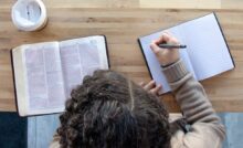 An overhead view shows a person sitting at a wooden table, writing in a notebook while reading the Bible.