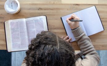 An overhead view shows a person sitting at a wooden table, writing in a notebook while reading the Bible.