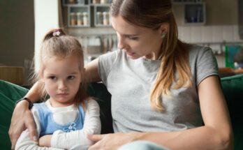 A mom and daughter sit on a green couch. The mom has her arm around her daughter, who has an upset expression.