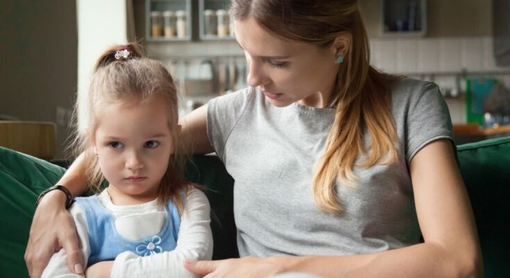 A mom and daughter sit on a green couch. The mom has her arm around her daughter, who has an upset expression.