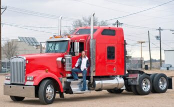 A female commercial truck driver standing on the step of her truck. The truck's cab is bright red and there's no trailer.