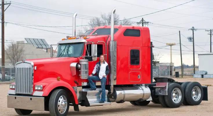 A female commercial truck driver standing on the step of her truck. The truck's cab is bright red and there's no trailer.