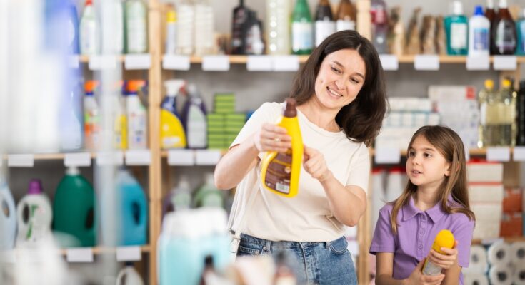 A mother and young daughter stand in a store aisle filled with cleaning supplies. The mother points to a can of cleaner.