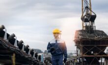 A man in a blue jumpsuit and yellow hardhat is holding a tablet and standing in the middle of an active mining site.