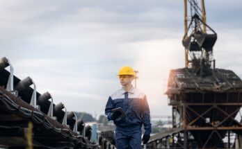 A man in a blue jumpsuit and yellow hardhat is holding a tablet and standing in the middle of an active mining site.