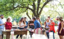 A group of people stand outside at a backyard barbecue. One man serves up food from a grill.