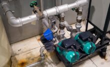 Two teal booster pumps are hooked up to metal pipes. The pumps sit on a black pedestal on a cream-colored tile floor.