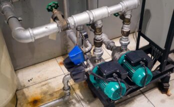 Two teal booster pumps are hooked up to metal pipes. The pumps sit on a black pedestal on a cream-colored tile floor.