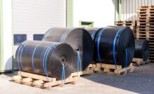 Multiple rolls of black plastic sheeting sit outside on top of wooden beams. They're secured with long blue and green cords.