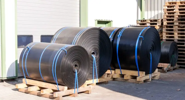 Multiple rolls of black plastic sheeting sit outside on top of wooden beams. They're secured with long blue and green cords.