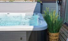 A white and brown swim spa has the top open and is full of water. A potted, tall, grassy plant sits next to the spa.
