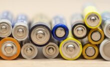 A stack of various sizes of batteries sits on a wooden surface. The batteries are blue, yellow, black, and silver.