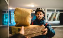 A delivery driver passes two pizza boxes and a bag to a person behind the camera. He smiles and wears a bike helmet.