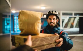 A delivery driver passes two pizza boxes and a bag to a person behind the camera. He smiles and wears a bike helmet.