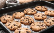 A darkly-colored tray contains four rows of three chocolate chip cookies. A white bowl of chocolate chips is next to the tray.