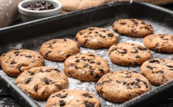 A darkly-colored tray contains four rows of three chocolate chip cookies. A white bowl of chocolate chips is next to the tray.