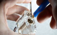 A close-up of a person using a tension wrench and a lock pick to manipulate the pins inside a clear practice lock.