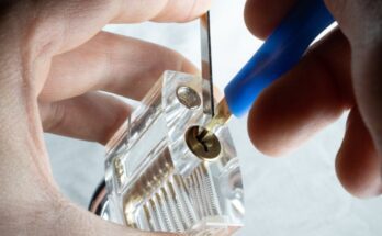 A close-up of a person using a tension wrench and a lock pick to manipulate the pins inside a clear practice lock.